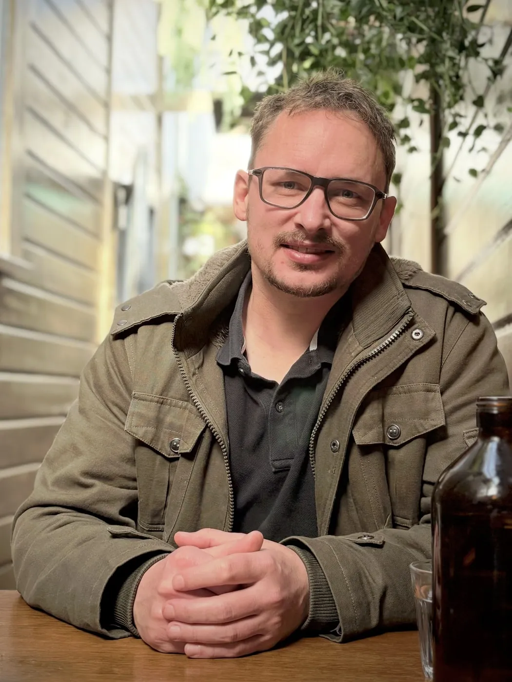 Weekday Phil at a café table, in glasses and an olive jacket